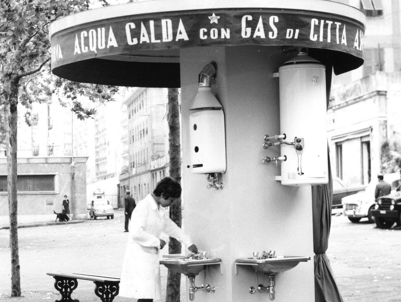 foto d'archivio, in bianco e nero, di una donna con camice bianco che apre un rubinetto di un distributore di acqua pubblica in strada