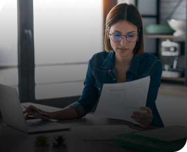 una donna seduta ad una scrivania, davanti a un pc e con dei fogli di carta che sta consultando