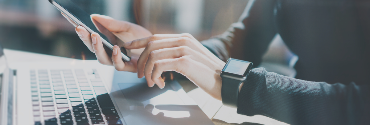 focus sulle mani di una donna con un cellulare in mano e sullo sfondo un pc