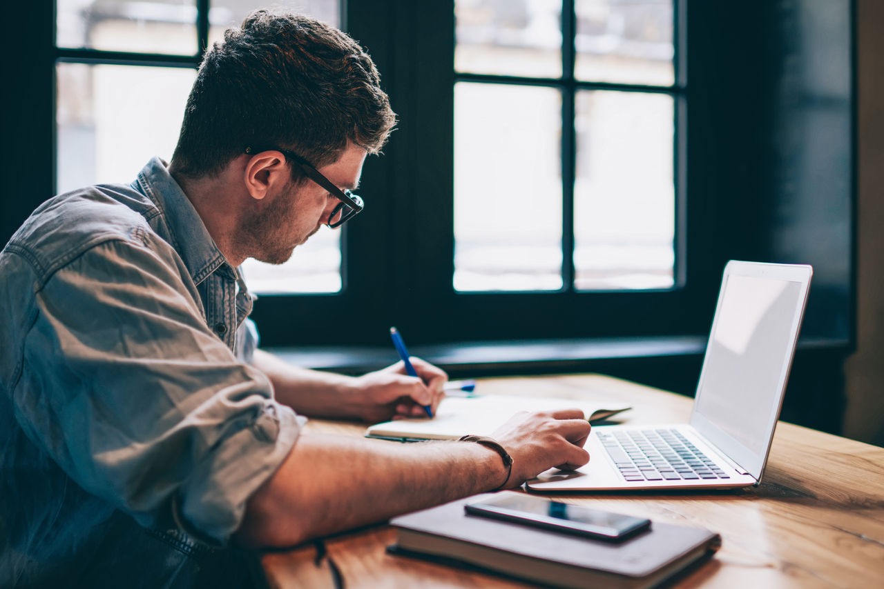 uomo che lavora alla scrivania davanti a un laptop, intento a scrivere su un quaderno